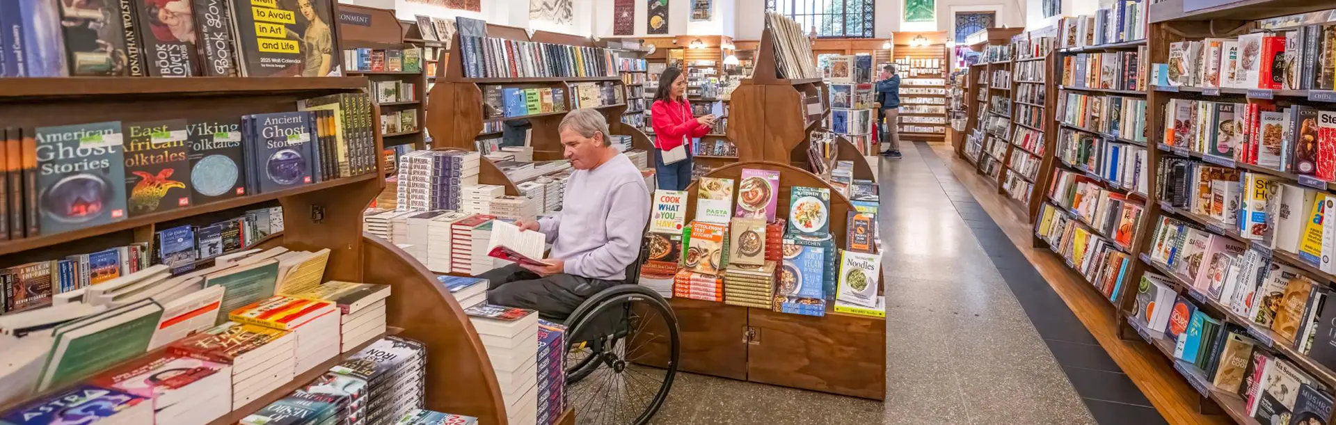 A man in a wheelchair and a woman in a pink coat shops for books in Munro's Books, a bookstore in Victoria, BC