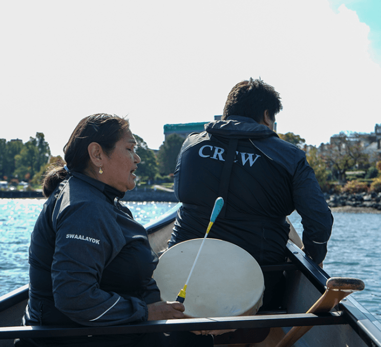 Two Indigenous people in a canoe, one is playing the drum