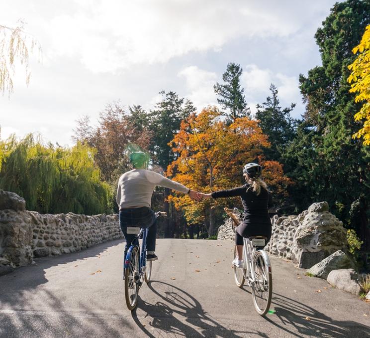 A couple rides bikes, hand-in-hand through Beacon Hill Park in Victoria, BC