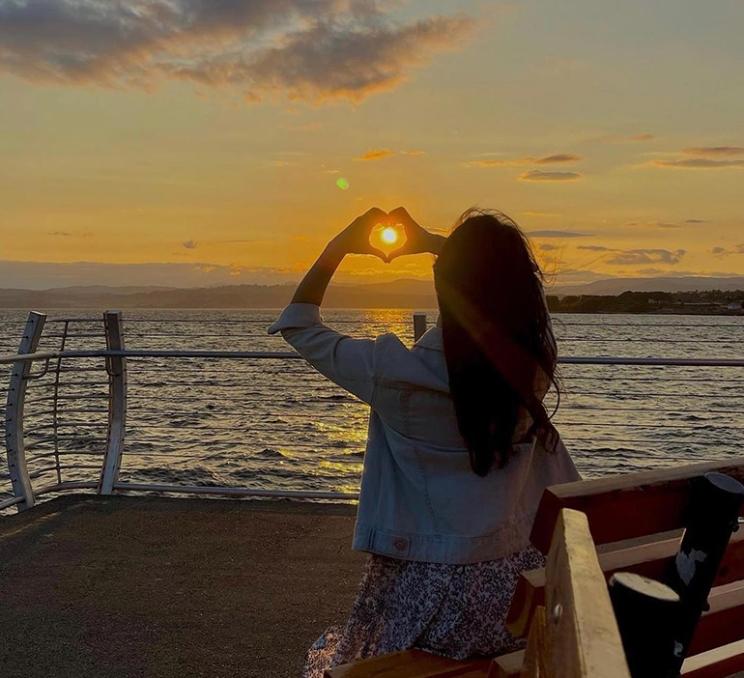 A woman makes a heart with her hands around the setting sun at the end of the Ogden Point Breakwater in Victoria, BC