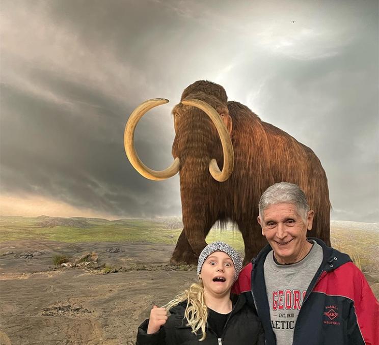 A grandfather and granddaughter take a photo in front of a wooly mammoth diorama at the Royal BC Museum in Victoria, BC