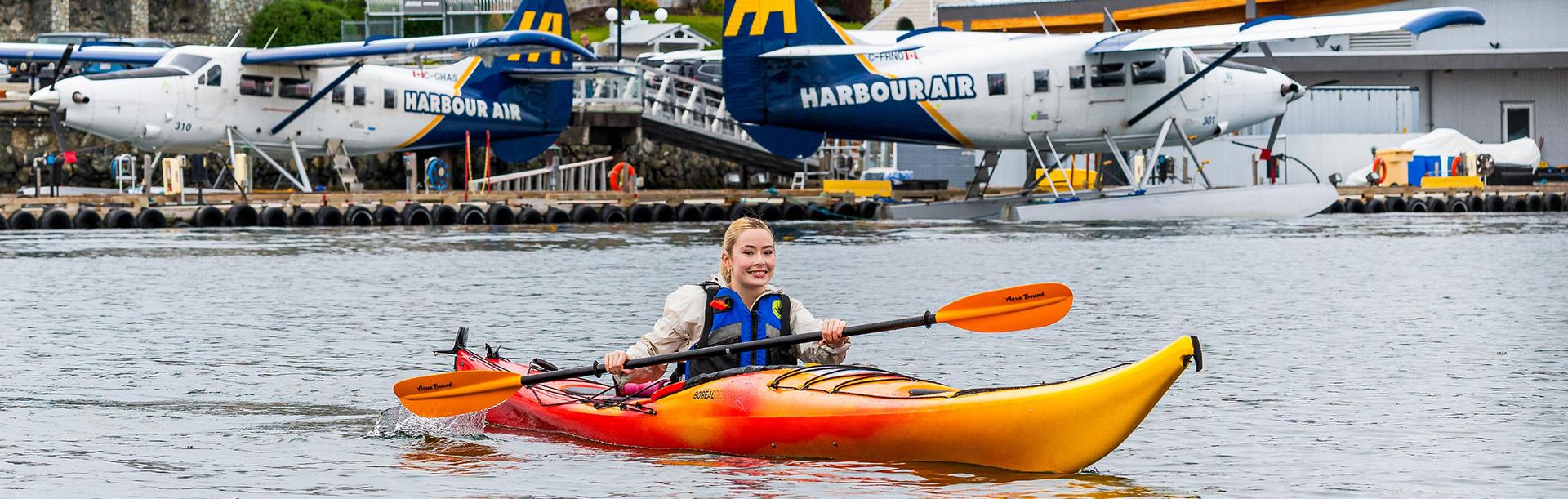 A kayaker paddles across the Inner Harbour in Victoria, BC with seaplanes moored along the dock behind her