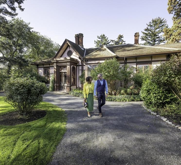 A woman in yellow and green and a man in blue walk down a pathway through the garden at Point Ellice House in Victoria, BC