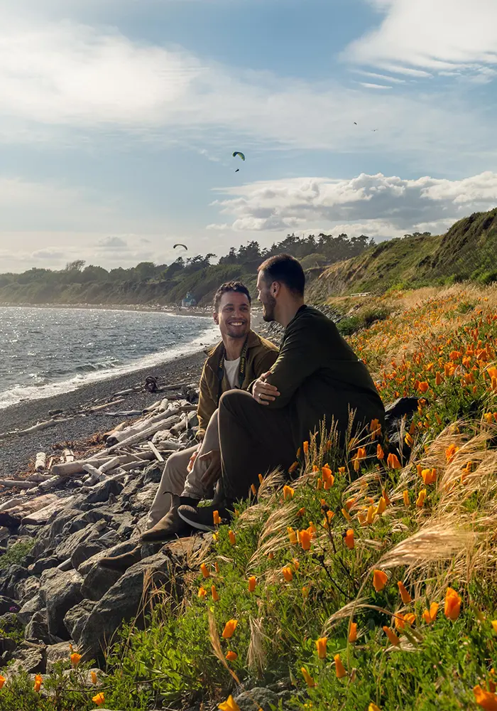 A male couple sits amidst wildflowers along the coast of Victoria, BC while paragliders sail through the sky behind