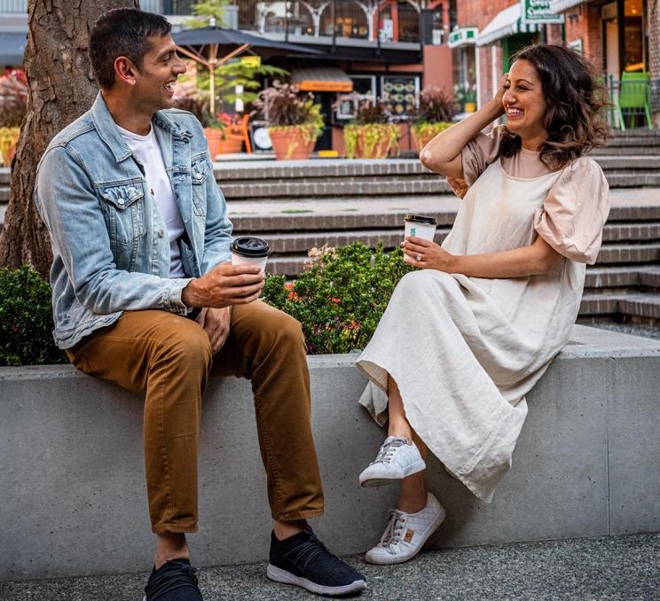A couple, a man dressed in a denim jacket and woman in a cream dress, enjoy a coffee in the courtyard of Market Square in Victoria, BC